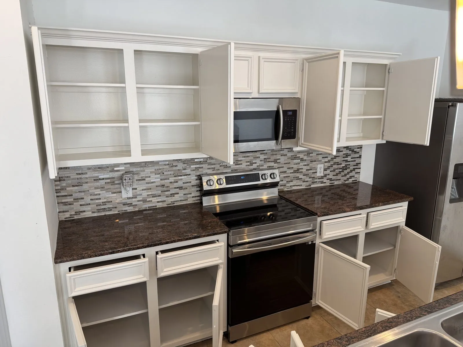 Kitchen cabinet boxes painted white with the doors still off, shelves empty and hinges exposed, dark granite counters and a mosaic backsplash behind