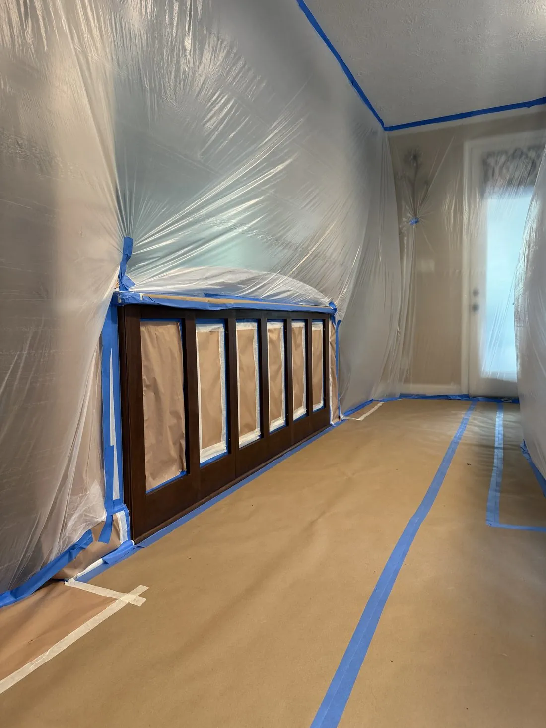 Long wall of kitchen cabinets with every door and drawer front removed, each opening papered and taped, the room tented in plastic and the floor covered