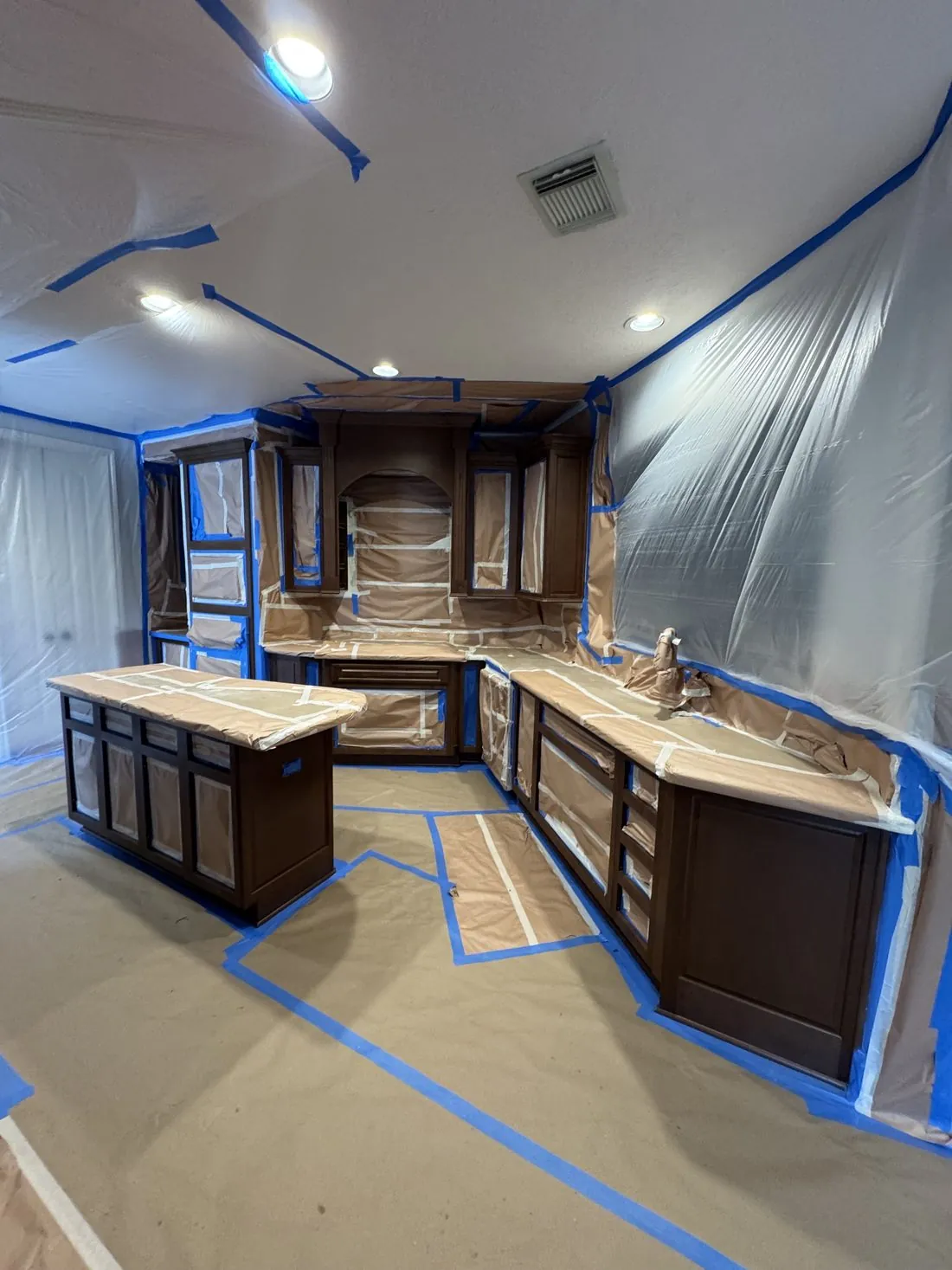Kitchen fully masked before cabinet painting, plastic sheeting taped floor to ceiling over the counters and appliances, blue tape lines on the papered floor and the dark cabinet boxes left exposed