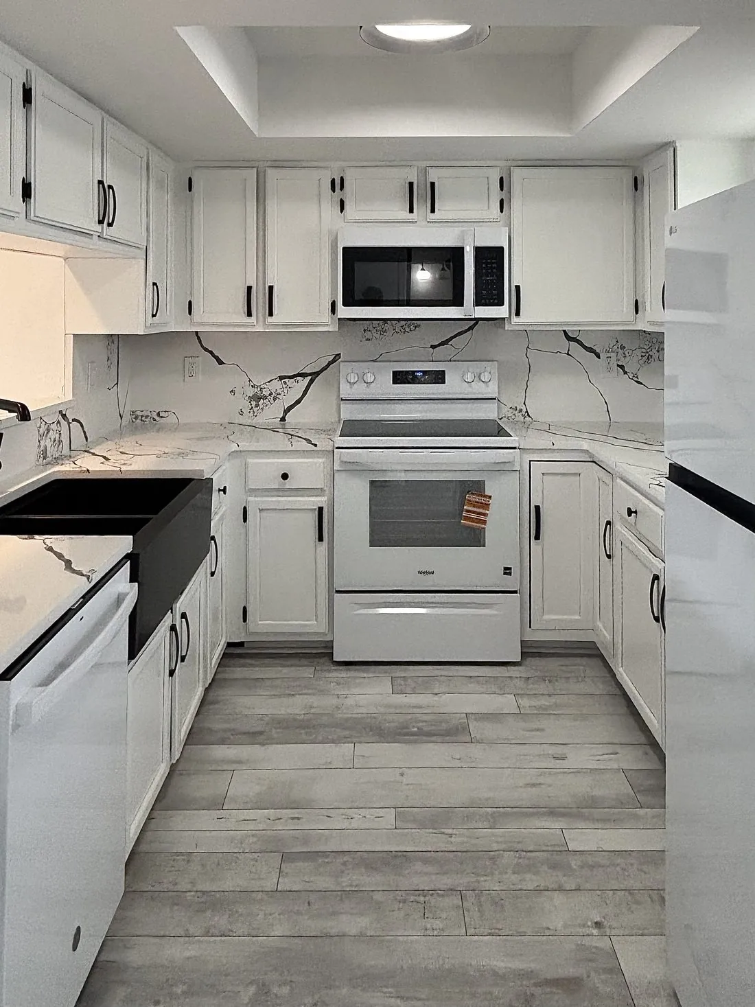 Kitchen after cabinet painting, white cabinets with black bar pulls under a tray ceiling, marbled counters and backsplash and gray plank flooring