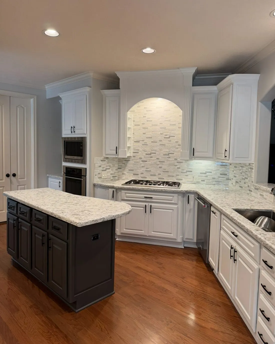 The same kitchen after painting, the perimeter cabinets and cooking arch now white with the island kept dark, granite counters and tile backsplash unchanged