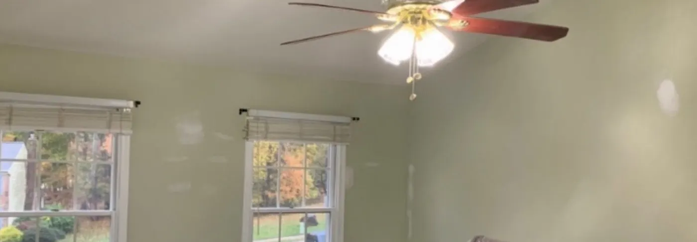 Bedroom before painting, pale sage green walls with a white chair rail, two windows and a ceiling fan, furniture under plastic