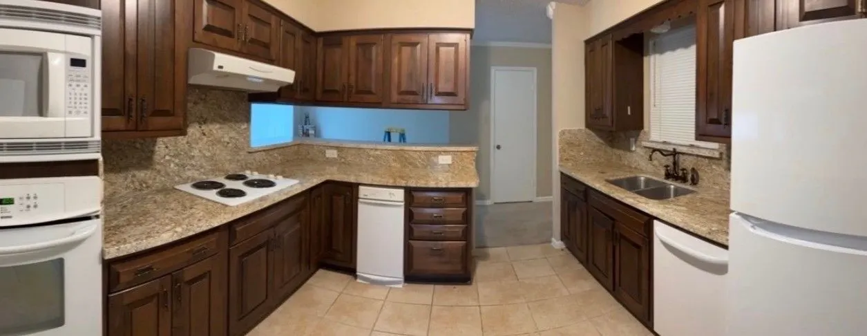 Galley kitchen before painting, dark stained wood cabinets above and below granite counters, white wall ovens and a white refrigerator