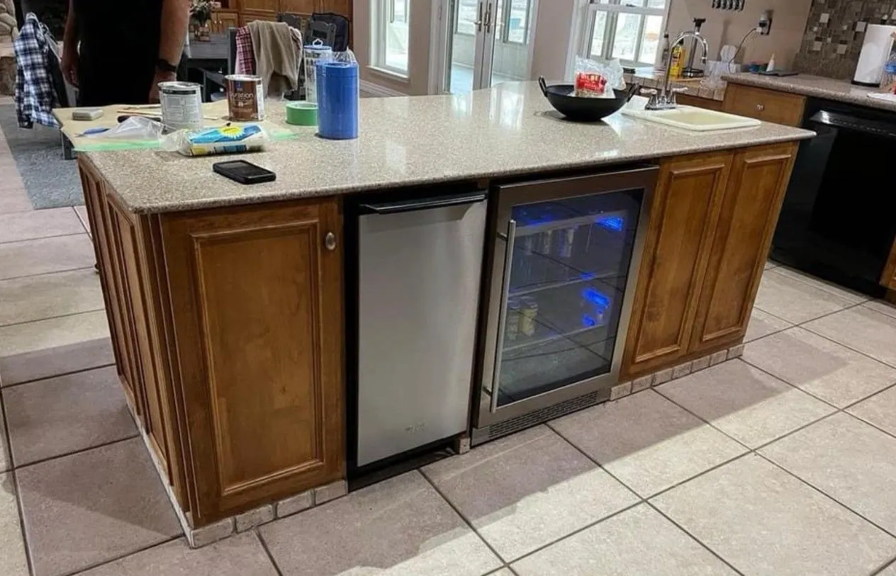 Kitchen island before painting, honey stained wood cabinet panels around a beverage fridge and a stainless dishwasher, tile floor