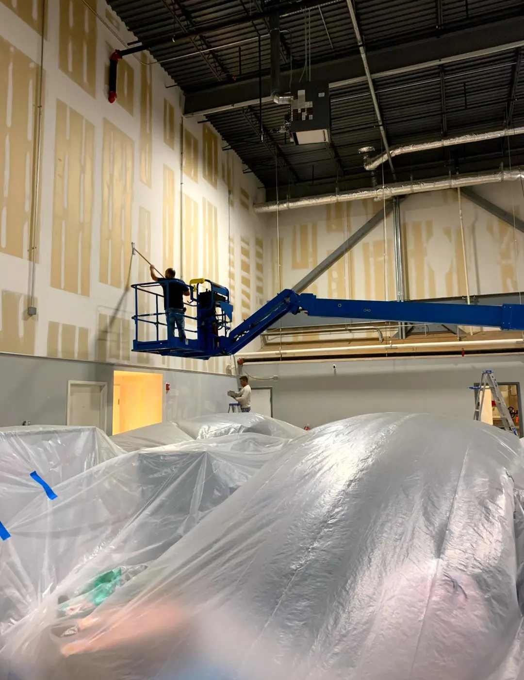 Crew painting the upper walls of a warehouse from a boom lift, the floor and everything below wrapped in plastic sheeting