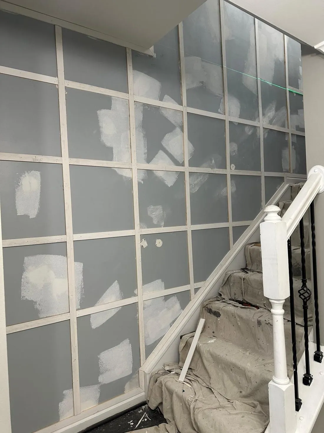 Stairwell accent wall before painting, a grid of applied molding on gray wall with white filler dabbed over every nail hole and joint