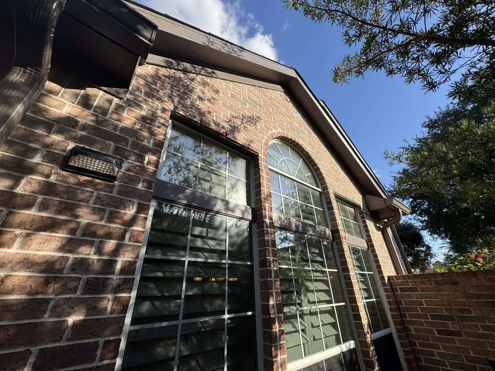 Tall arched window on a red brick home, the surrounding trim and the eave above it repainted dark brown, photographed from below