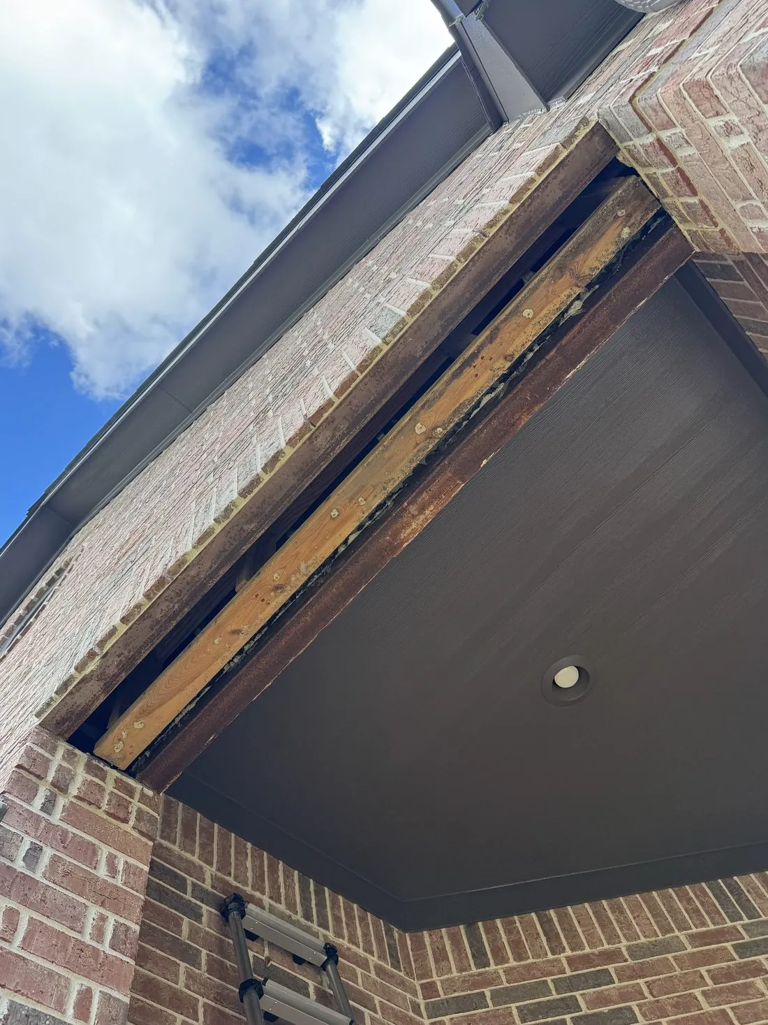 Eave on a brick home mid-job, the rotted fascia board removed to expose bare rafter tails while the soffit above it is already painted dark brown