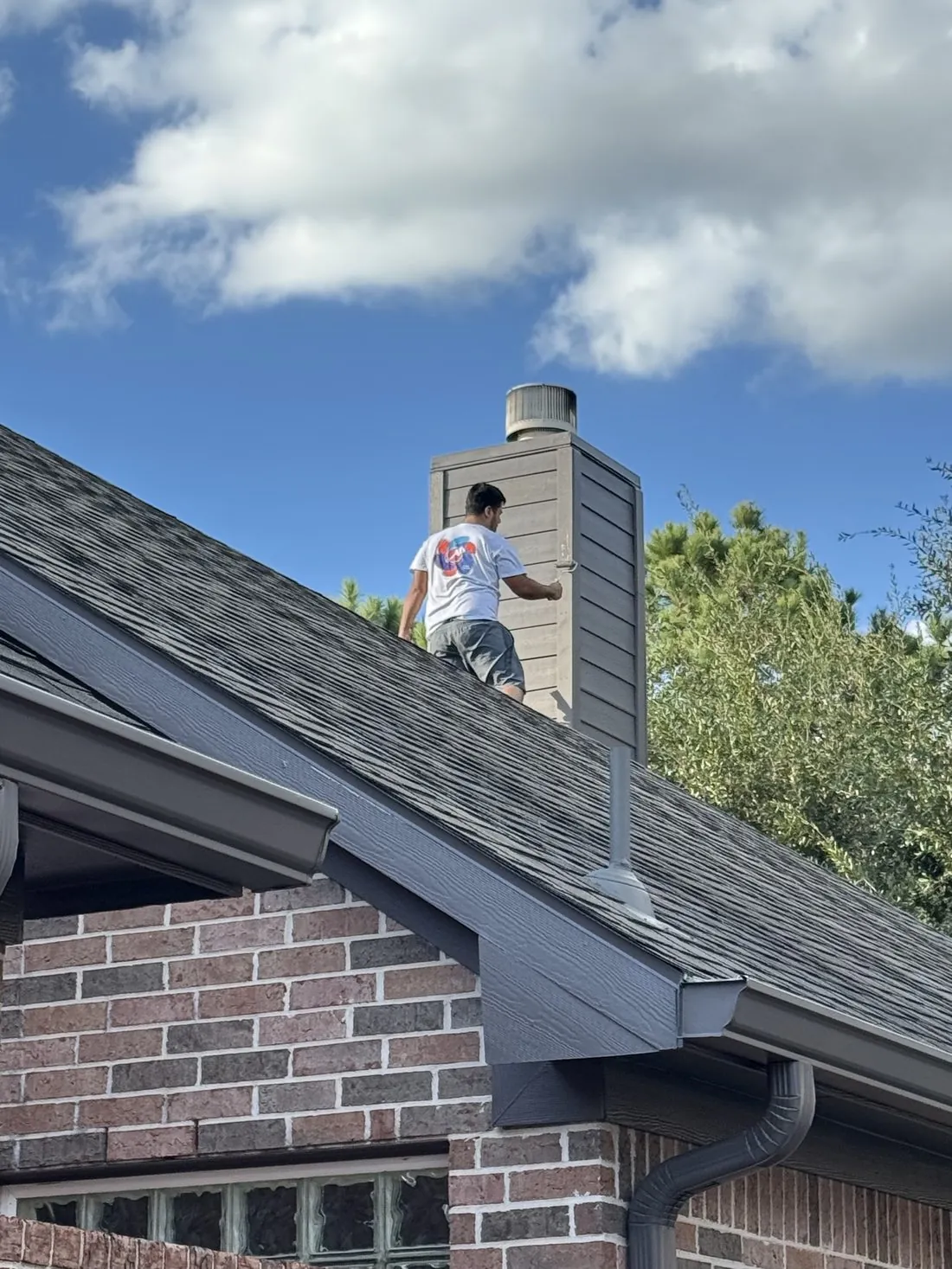Painter standing on the shingle roof of a brick home beside the chimney chase, working on the siding above the gutter line