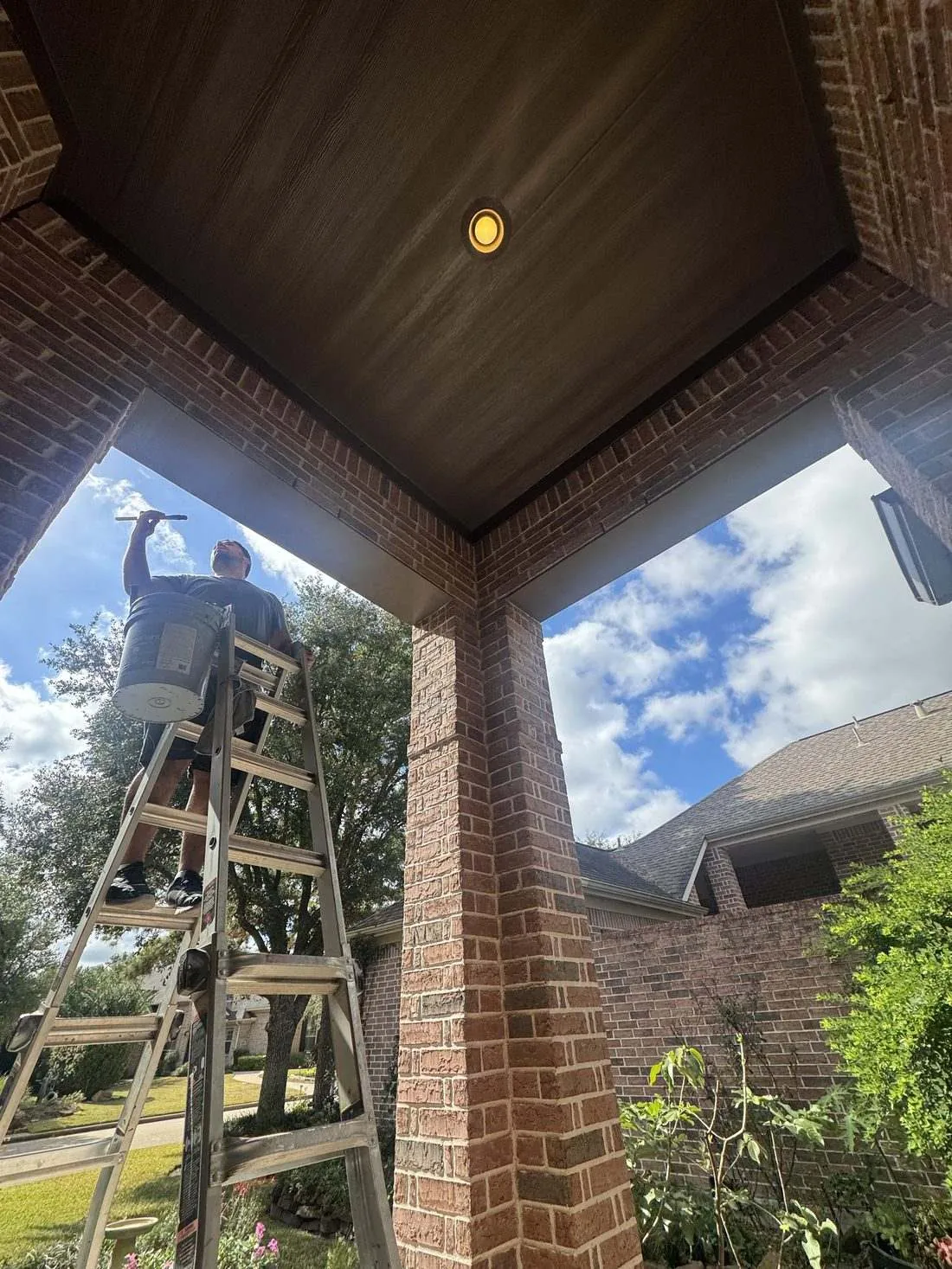 Painter on a wooden stepladder under the covered porch of a brick home, holding a bucket and cutting in the dark brown porch ceiling by hand